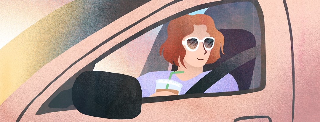 In a car, a woman with an iced coffee in hand looks out of the driver side window at the sunrise.